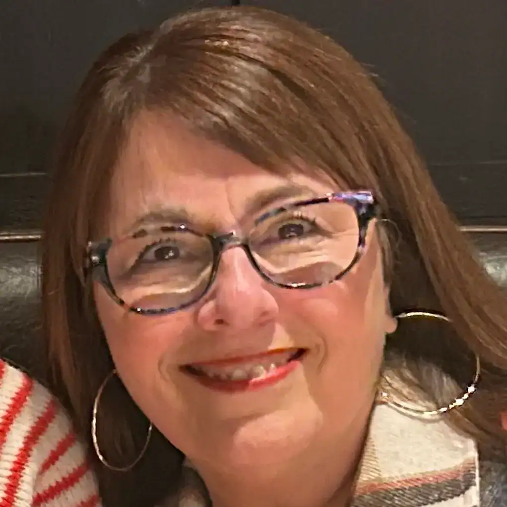 Smiling woman with glasses, hoop earrings, and shoulder-length brown hair, seated indoors.